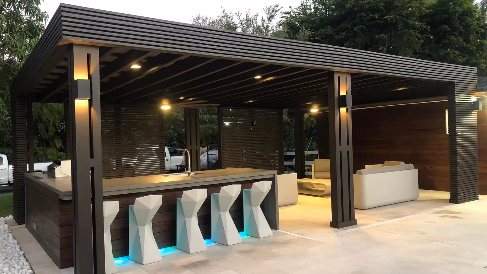 Dark wood pavilion with outdoor bar, sculptural stools, LED toe-kick lights and lounge at dusk.