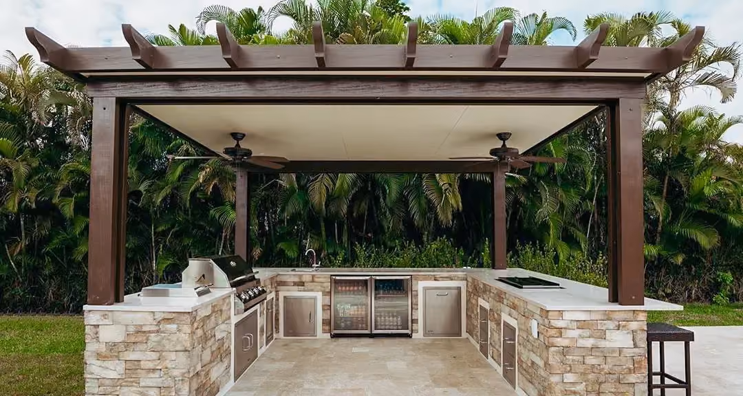 Brown pergola with two ceiling fans over a U-shaped stacked stone outdoor kitchen.