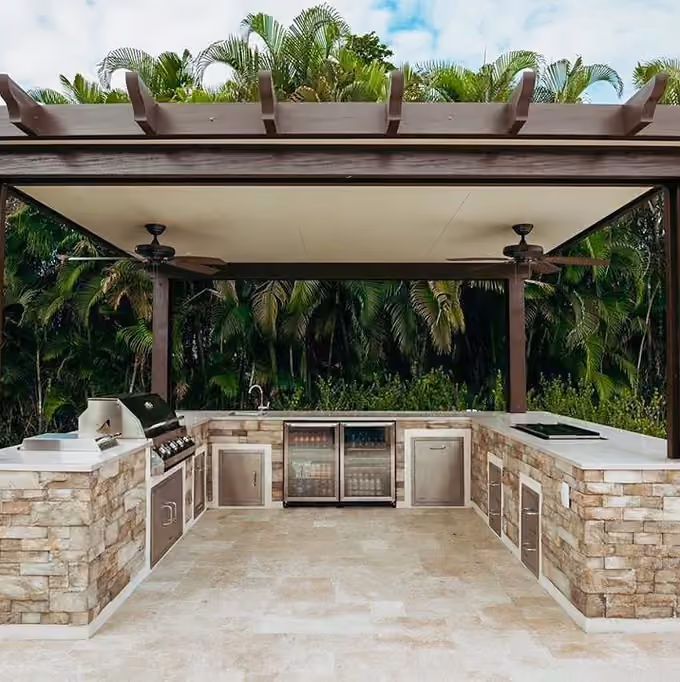 Brown pergola with two ceiling fans over a U-shaped stacked stone outdoor kitchen.