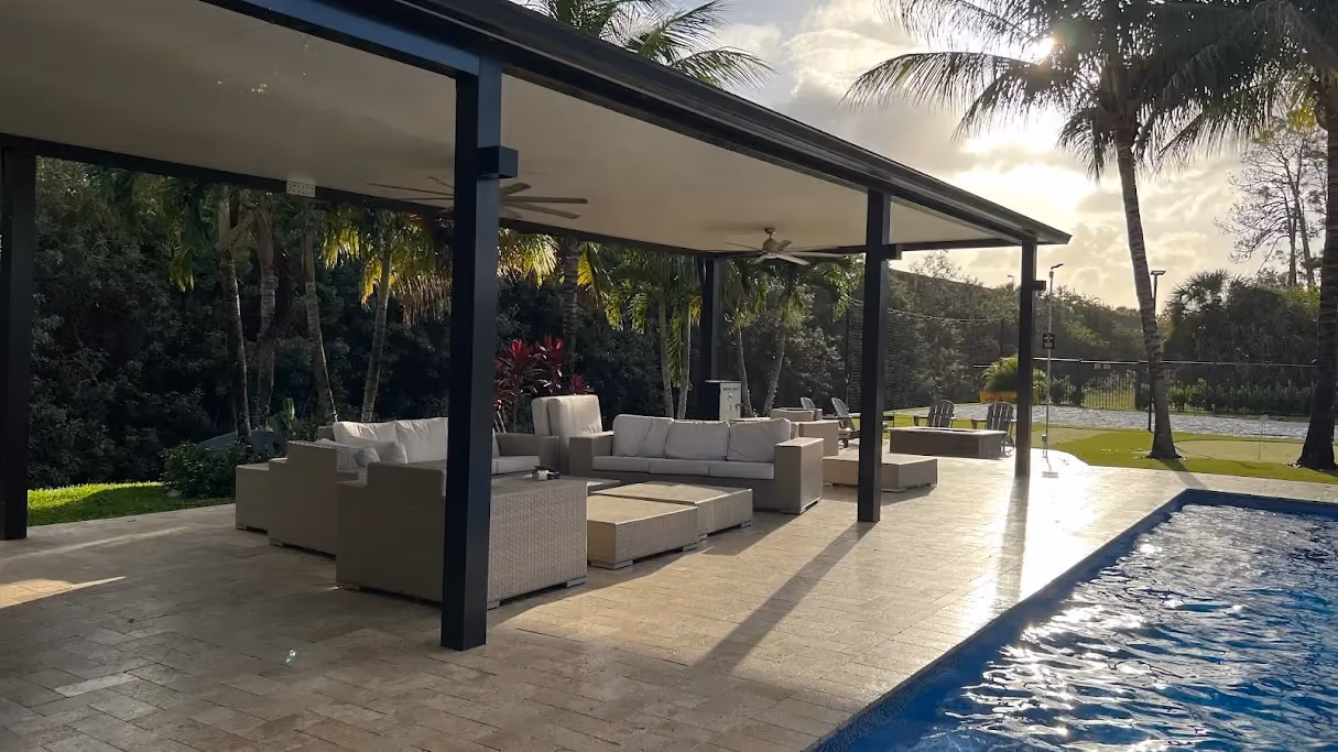 Black-framed insulated patio cover with ceiling fans and wicker sofas beside a lap pool at sunset