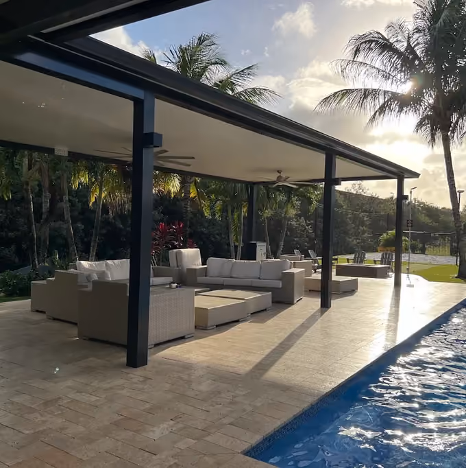 Black-framed insulated patio cover with ceiling fans and wicker sofas beside a lap pool at sunset