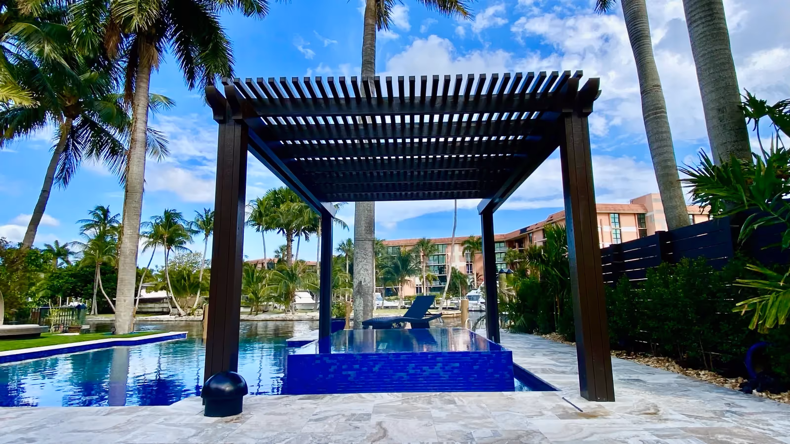 Dark bronze freestanding lattice pergola framing a blue-tiled spa with canal, boats and palms behind