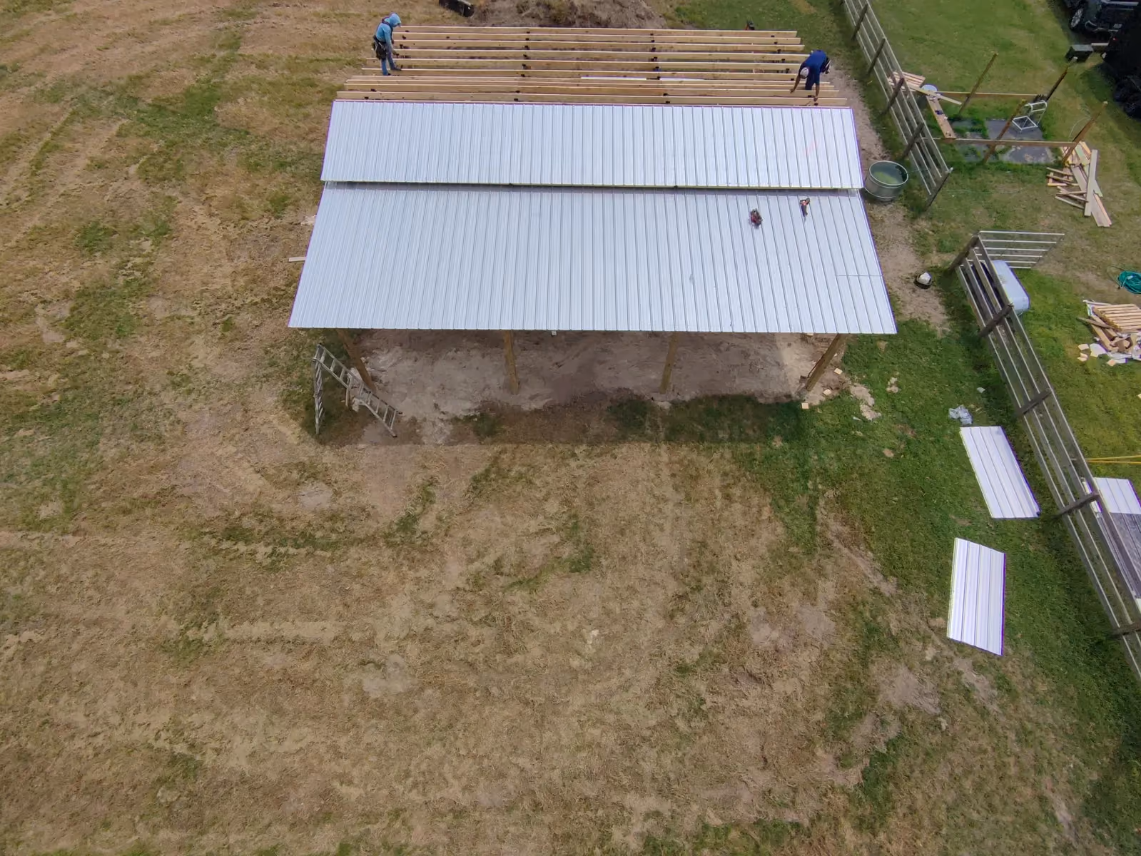 Aerial view of galvanized metal roof panels being installed over the purlins of a pole barn.