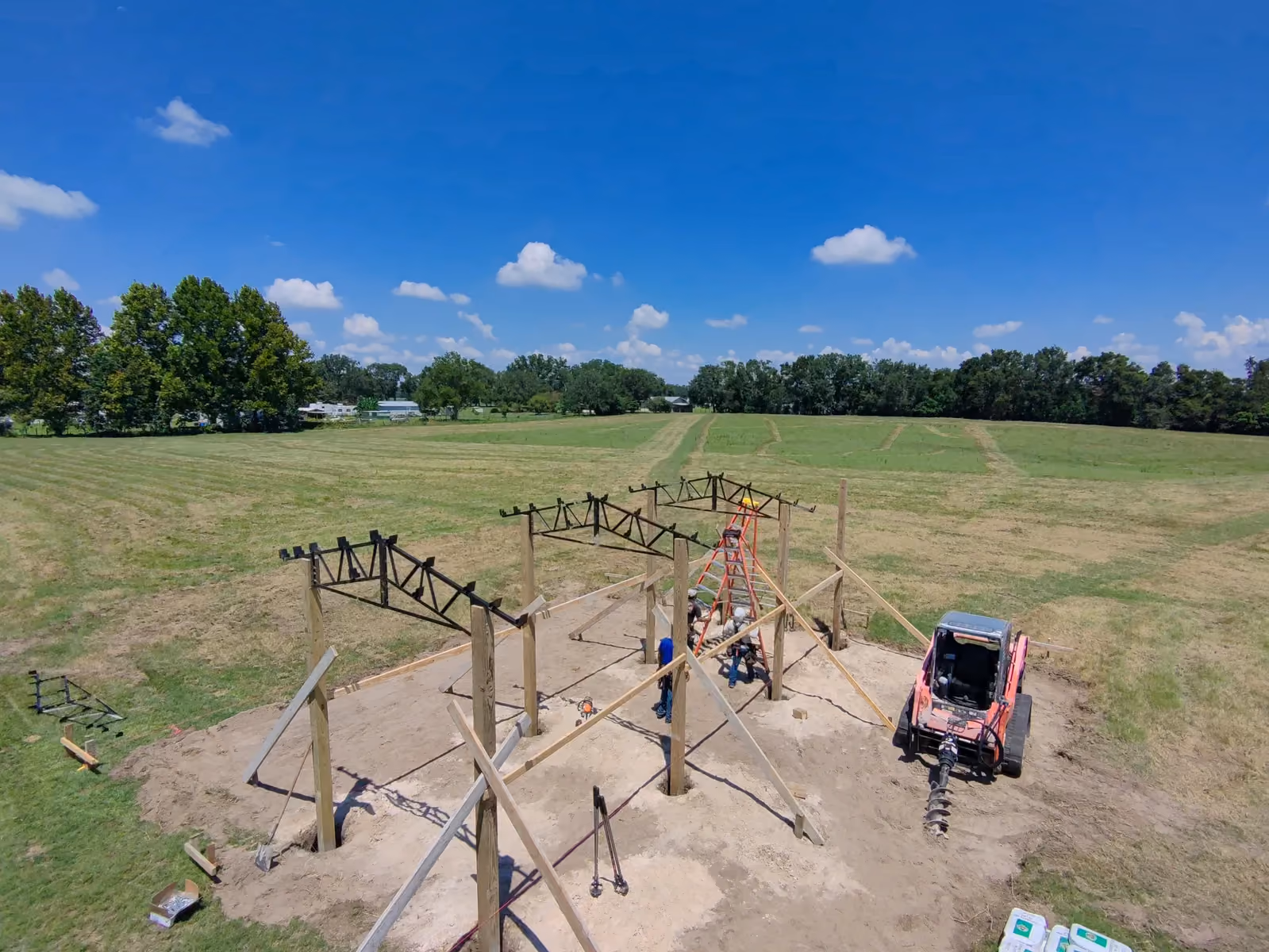 Aerial view of pole barn posts with three steel trusses set, braced with lumber, and an auger skid steer.