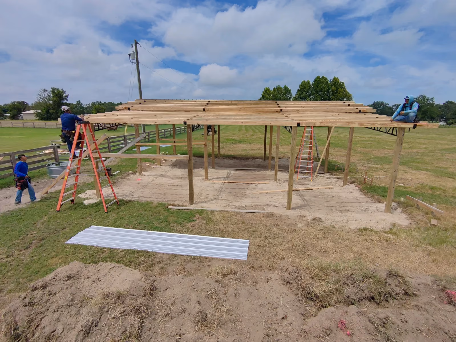 Crew installing wood roof purlins on a pole barn frame, with metal roofing panels staged on the grass.