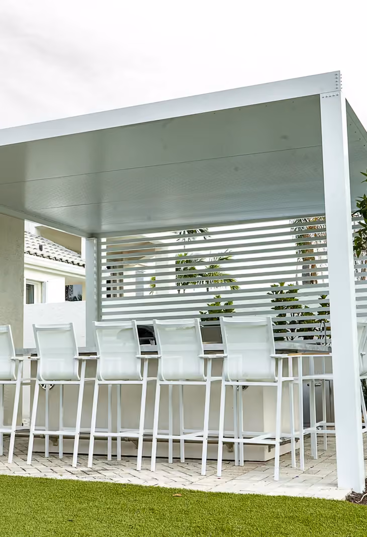 White aluminum pergola with a slatted privacy wall and a row of white bar stools on a paver patio.