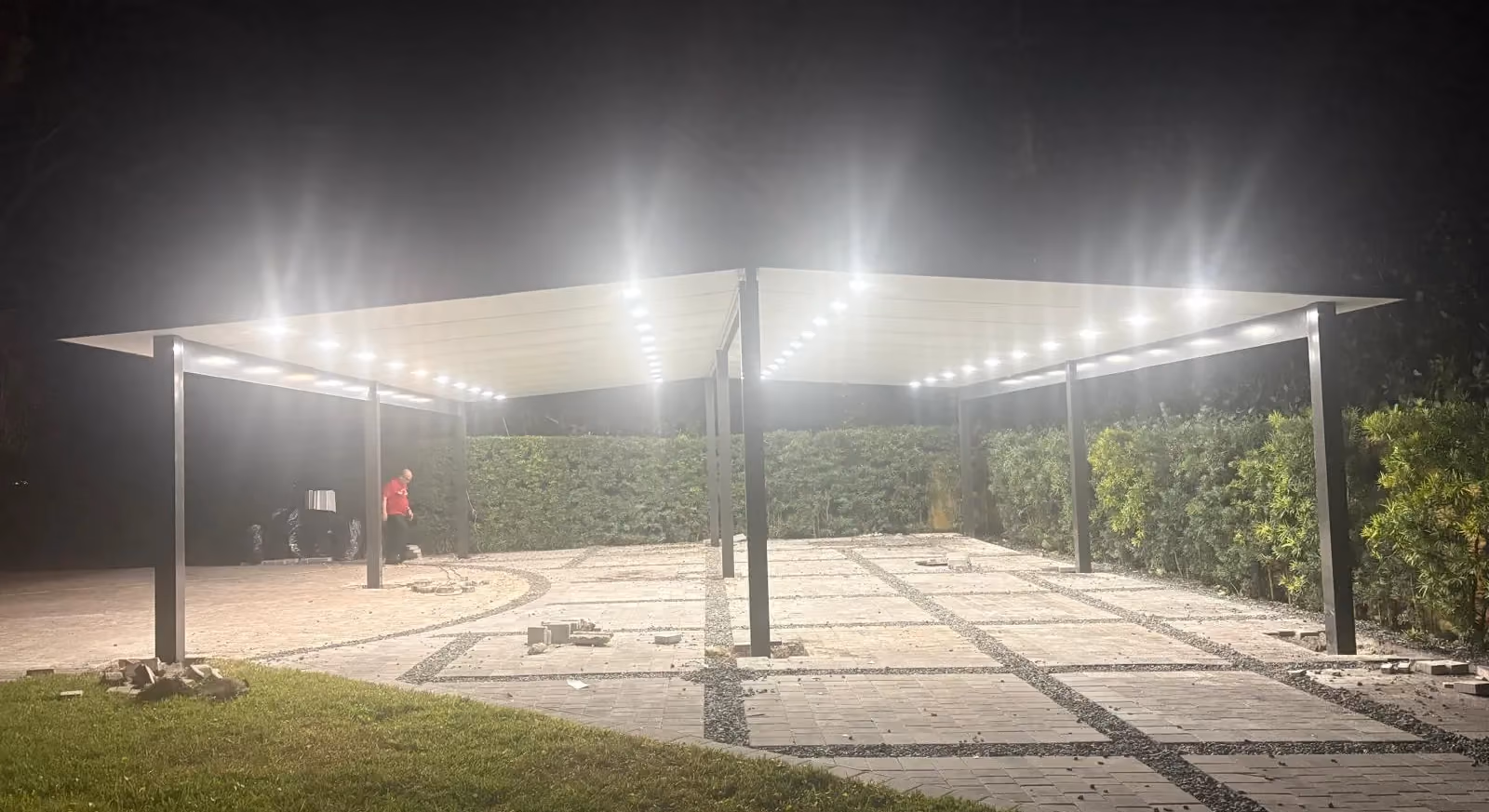 Large dark-framed insulated patio cover with rows of recessed lights glowing at night over a paver court