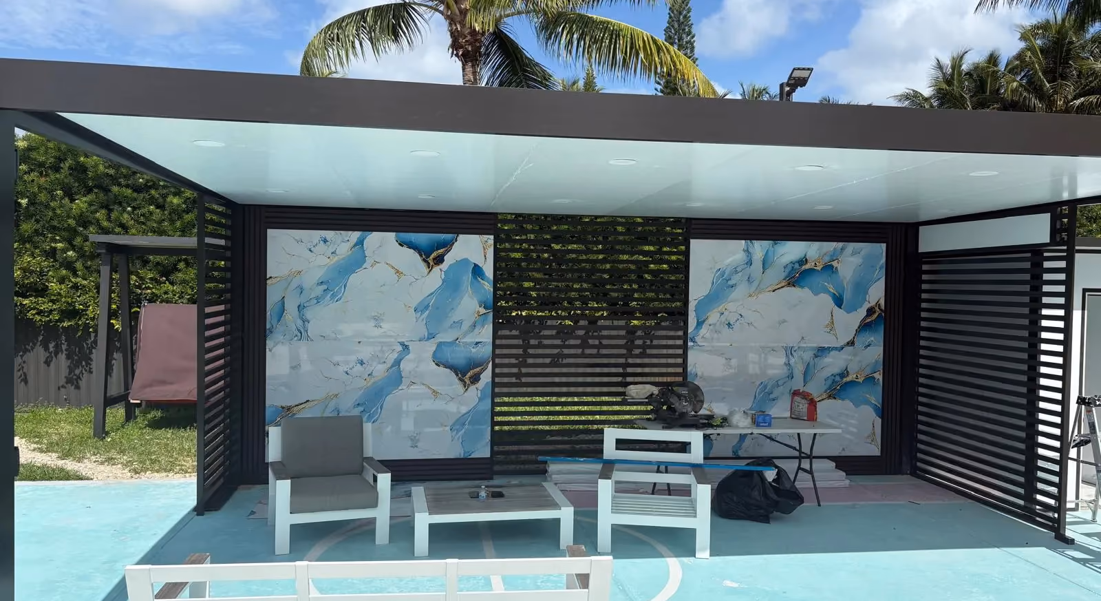 Black-framed flat patio cover with slatted walls and blue marble-look panels being installed over a sports court