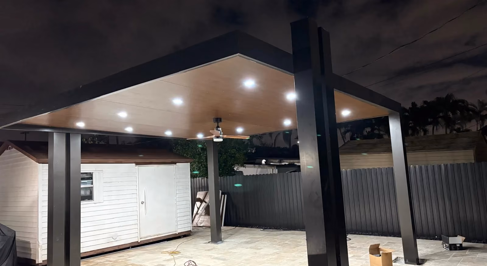 Black-framed patio cover with woodgrain ceiling, recessed lights and fan at night over a travertine patio