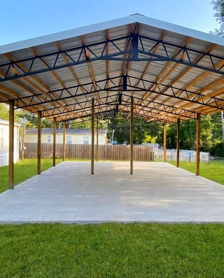 Open pole barn with a metal gable roof and black trusses over a new concrete slab.