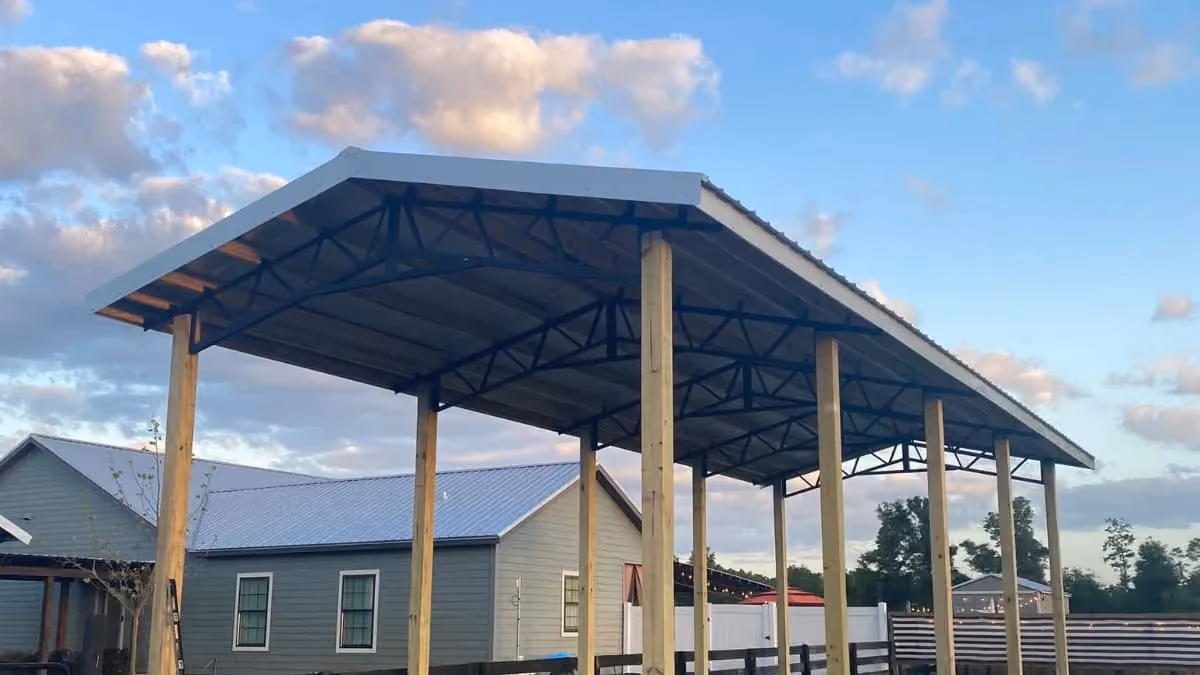 Narrow open pole barn with new wood posts, black steel trusses and metal roof beside a farmhouse.