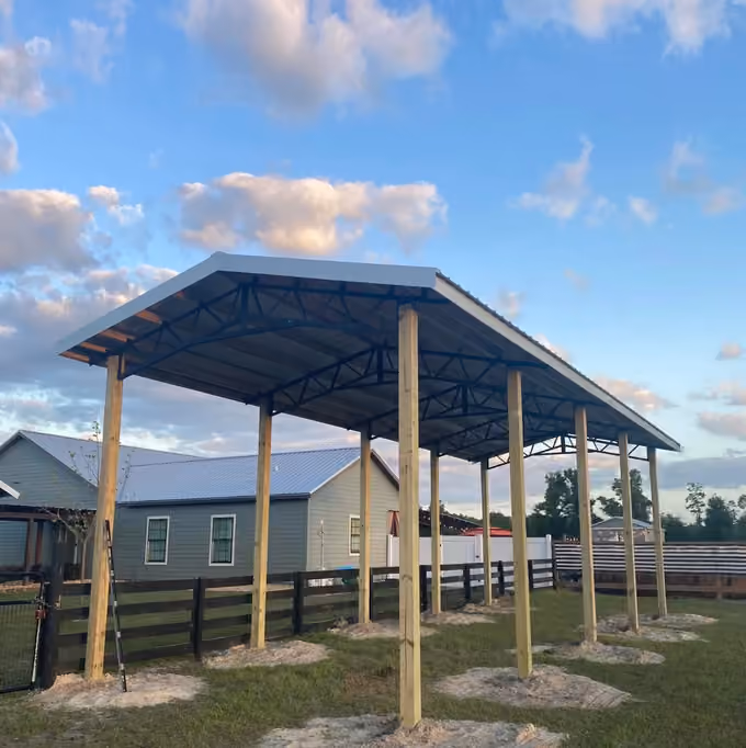 Narrow open pole barn with new wood posts, black steel trusses and metal roof beside a farmhouse.
