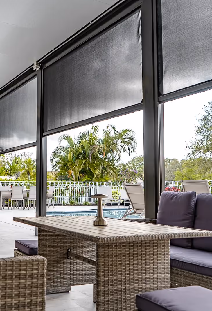 Motorized retractable screens half lowered on a covered patio, with wicker seating and palms beyond.