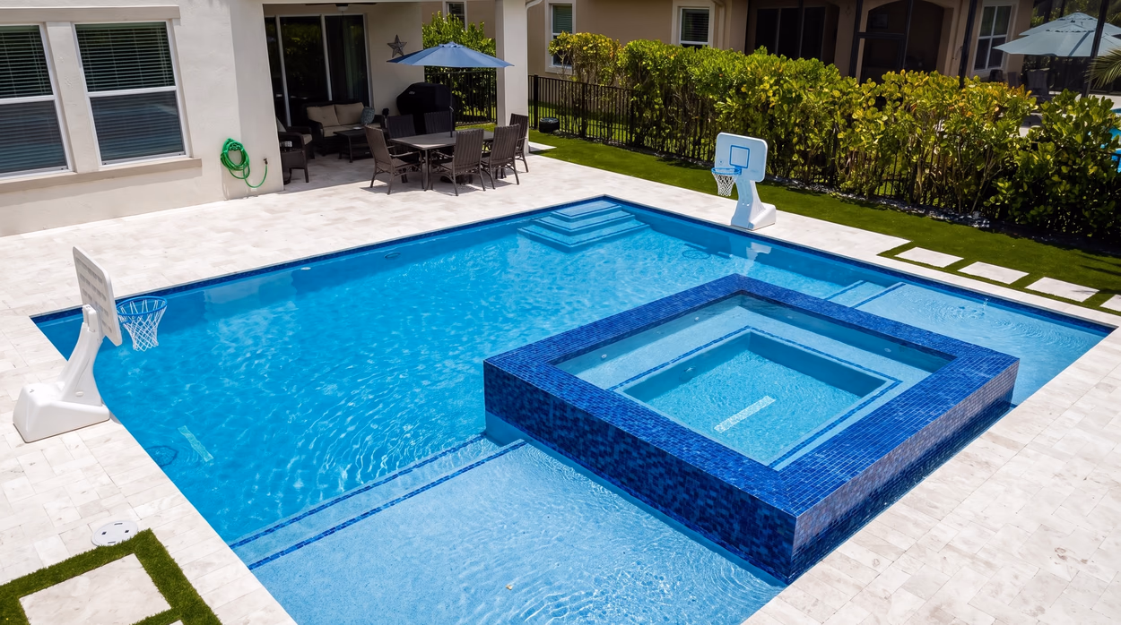 Rectangular pool with a raised blue glass mosaic spa spilling into the water, framed by a travertine paver deck in a South Florida backyard.