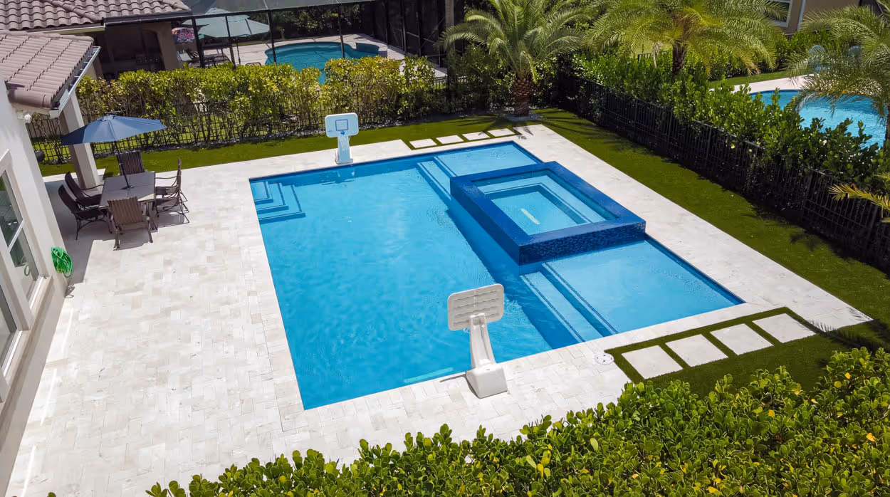 Modern rectangular backyard pool with attached spa, basketball hoops, surrounded by white tile and green landscaping.