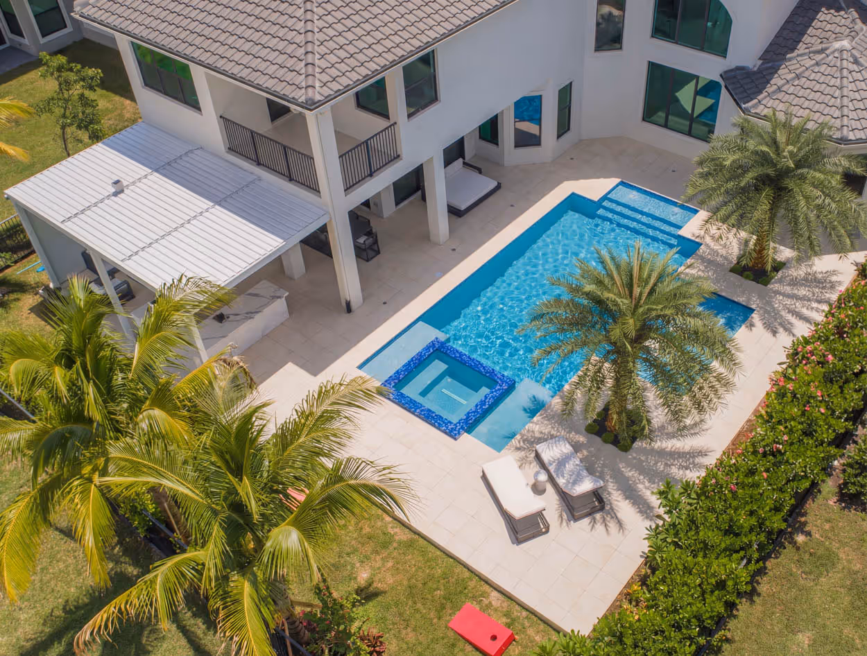 Aerial view of a modern backyard with a rectangular swimming pool, attached spa, palm trees, lounge chairs on the patio, and a two-story house with a tiled roof.